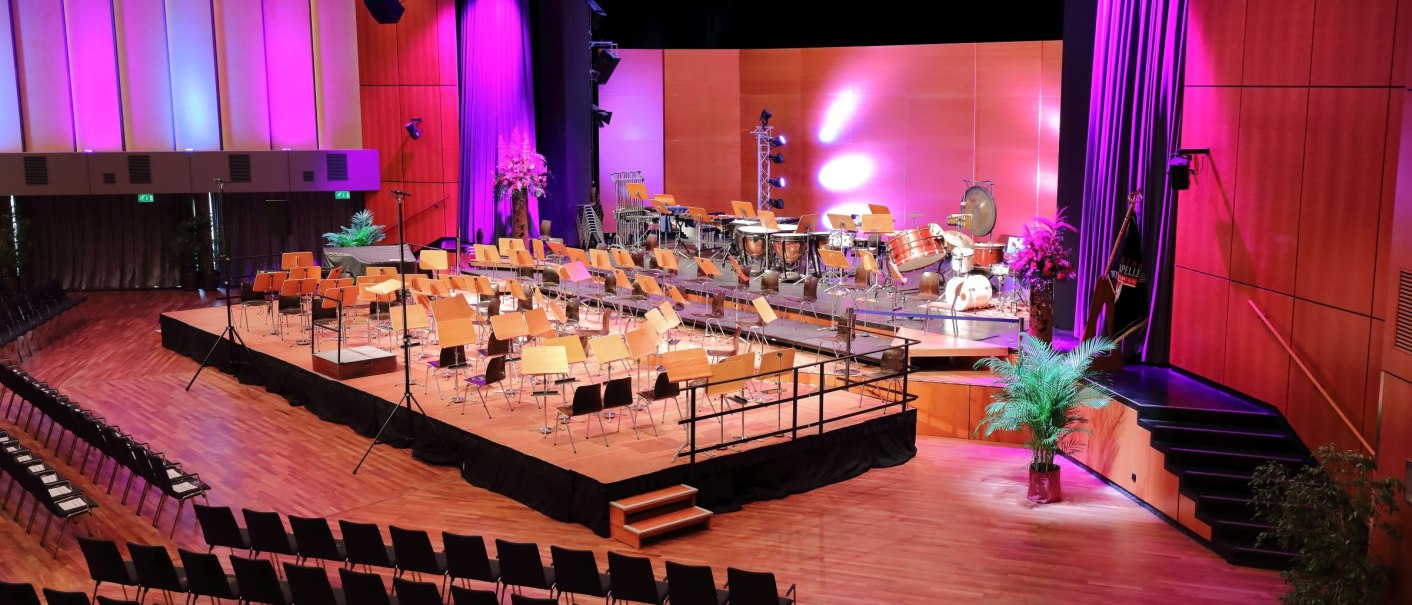 Concert hall with empty chairs and instruments on the stage, purple lighting, wooden floor, plants on the sides., © ccbs-gf