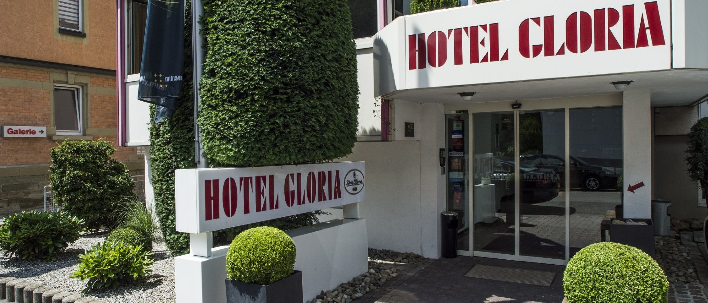 Entrance to Hotel Gloria with large sign and well-tended plants. A modern glass front and decorative shrubs frame the entrance., &copy; Hotel Gloria