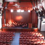 An old gymnasium with wooden beams, a stage and many rows of chairs. The stage is equipped with curtains and a lectern., © Stadt Herrenberg