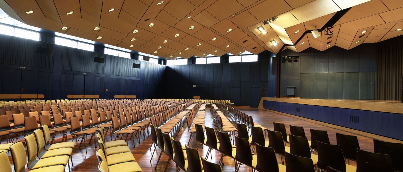 Großer Saal der Osterfeldhalle mit leeren Stuhlreihen, einer Bühne und einer modernen Deckenstruktur., © eslive Großer Saal der Osterfeldhalle mit leeren Stuhlreihen, einer Bühne und einer modernen Deckenstruktur., © eslive