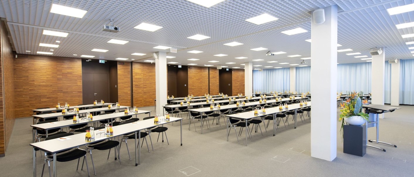 Großer Konferenzraum mit Tischen und Stühlen, Flaschen und Gläsern auf den Tischen. Holzverkleidung an den Wänden, helle Deckenbeleuchtung., © Geno-Haus