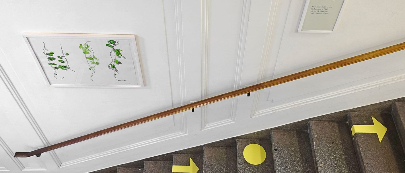Treppenhaus mit gelben Pfeilen und Abstandsmarkierungen. An der Wand hängen Bilder und ein Handlauf verläuft parallel zur Treppe., © Literaturhaus Stuttgart