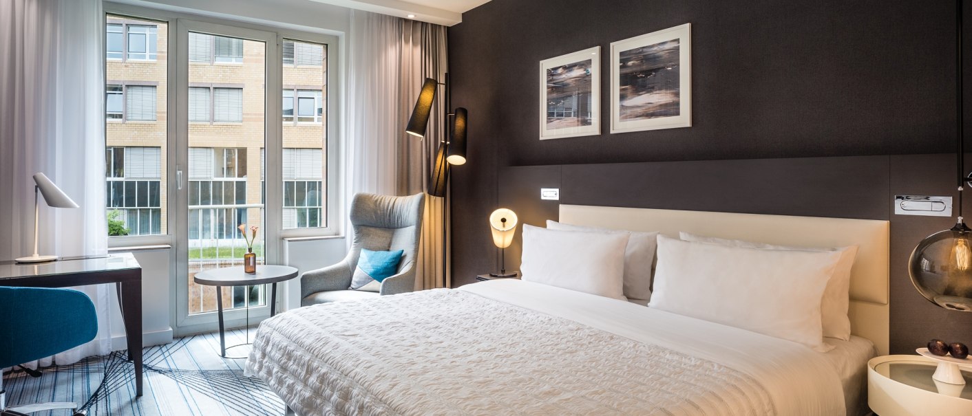 Modernes Hotelzimmer mit gro&szlig;em Bett, Schreibtisch, Sessel und Fenster mit Blick auf ein Geb&auml;ude., &copy; Le M&eacute;ridien Stuttgart, Gerrit Meier