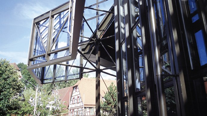 A modern glass facade with geometric patterns, with traditional half-timbered houses and trees in the background under a blue sky., &copy; Stadt Sindelfingen