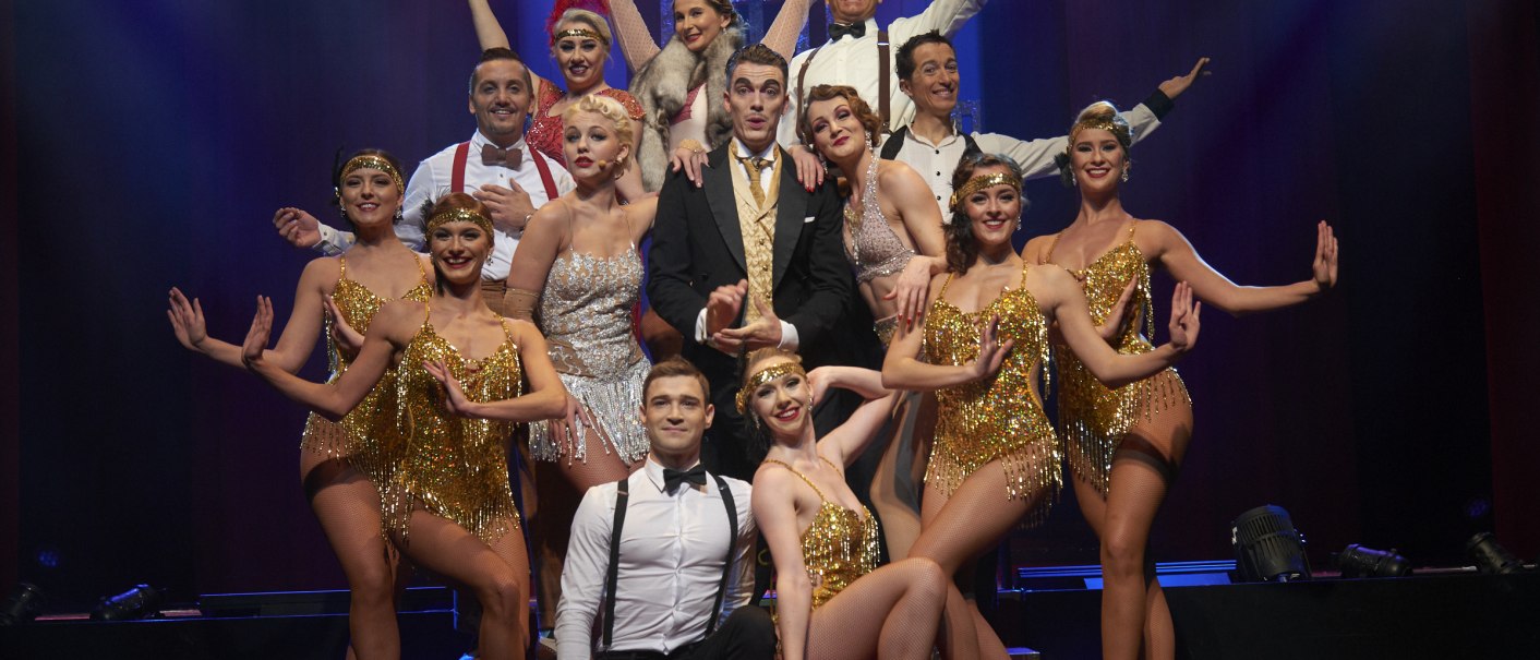 The Friedrichsbau Varieté Ensemble poses on stage in glittering costumes, surrounded by colorful lights., © Alexandra Klein