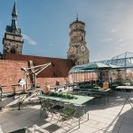 Dachterrasse mit gr&uuml;nen Tischen und St&uuml;hlen, umgeben von Glasstrukturen. Im Hintergrund sind zwei Kircht&uuml;rme zu sehen, unter blauem Himmel., &copy; OutOfOffice GmbH