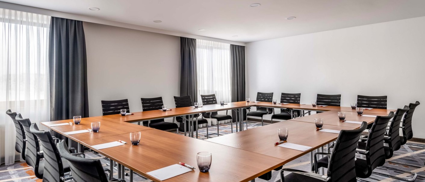 Modern meeting room with U-shaped table arrangement, black chairs, notepads and glasses. Large windows with curtains let in daylight., &copy; copyright by Kilian Blees