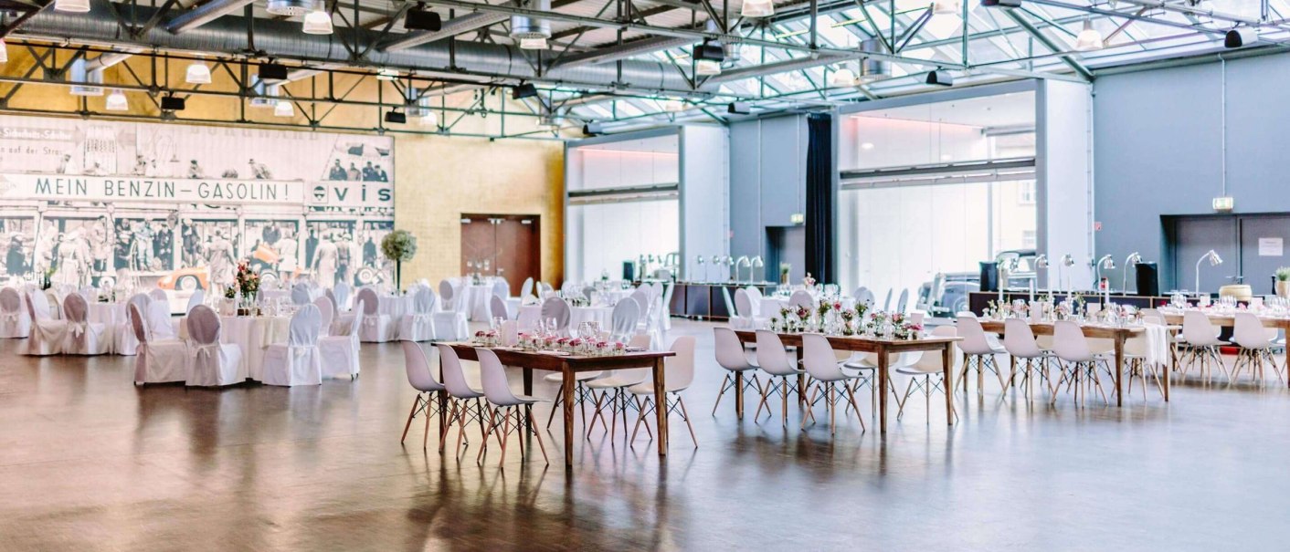 Großer, heller Raum mit gedeckten Tischen und Stühlen, dekoriert für ein Event. Im Hintergrund ein Wandbild mit historischem Motiv., © rauschenbergercatering