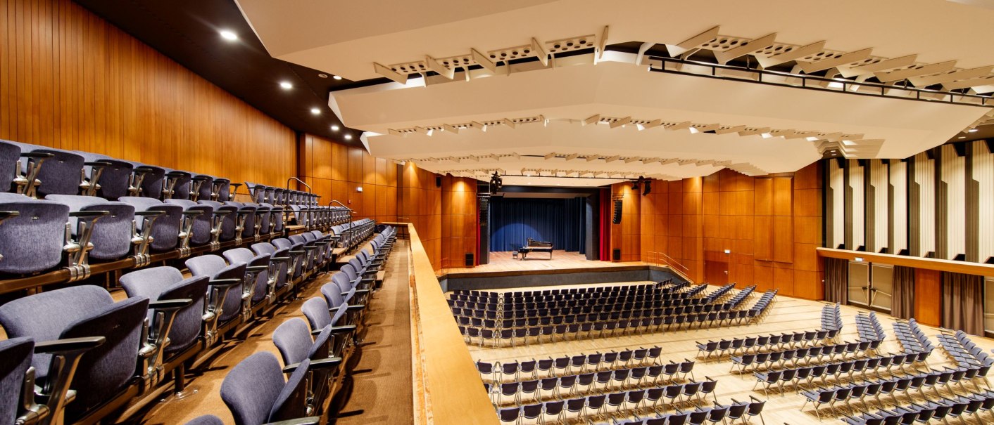 Großer Saal der Stadthalle Göppingen mit leeren blauen Sitzreihen, Holzwänden und einer Bühne mit Flügel., © Tobias Fröhner Photography