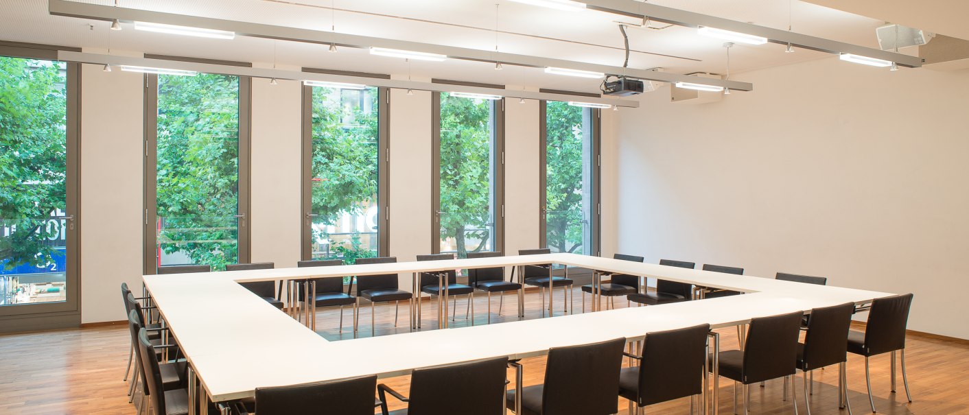Moderner Konferenzraum mit U-förmigem Tisch und schwarzen Stühlen, große Fenster mit Blick auf grüne Bäume., © hdkk_tf
