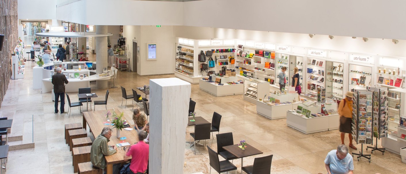 Modernes Atrium mit Sitzbereichen, einem Shop und Menschen, die sich unterhalten. Helle, offene Architektur mit Glasgeländer und Steinboden., © hdkk_gf