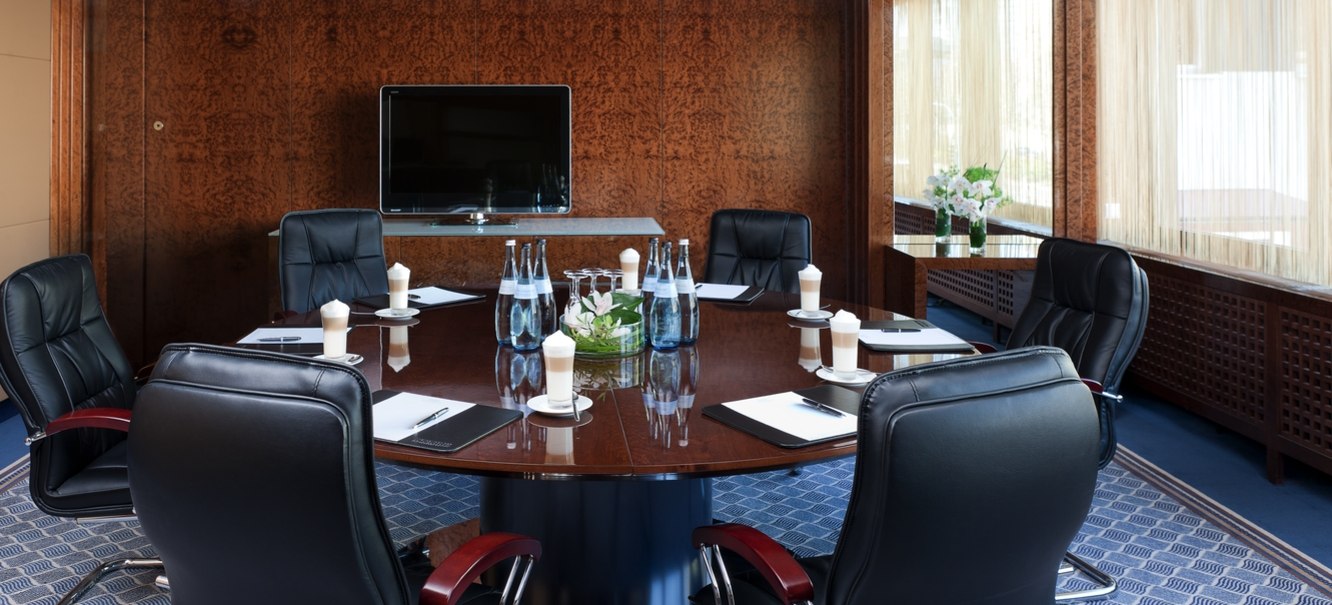 Eleganter Konferenzraum mit rundem Holztisch, Ledersesseln, TV an der Wand und Fenstern mit Vorhängen. Auf dem Tisch stehen Wasserflaschen und Notizblöcke., © Steigenberger Hotels AG Eleganter Konferenzraum mit rundem Holztisch, Ledersesseln, TV an der Wand und Fenstern mit Vorhängen. Auf dem Tisch stehen Wasserflaschen und Notizblöcke., © Steigenberger Hotels AG