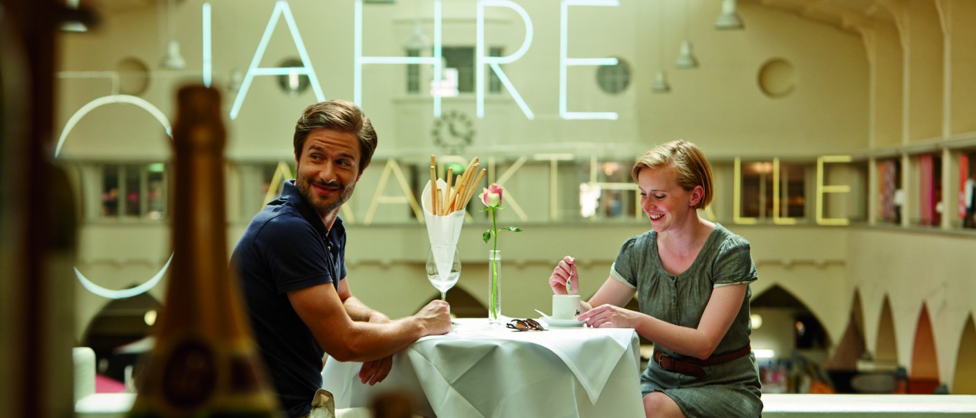 A couple sits smiling at a table in a café. Large letters are visible on the wall in the background., © TMBW, Christoph Düpper A couple sits smiling at a table in a café. Large letters are visible on the wall in the background., © TMBW, Christoph Düpper