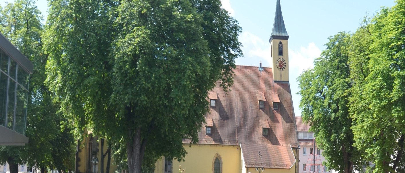 Die Kreuzkirche N&uuml;rtingen mit ihrem markanten Turm, umgeben von gr&uuml;nen B&auml;umen und roten Sonnenschirmen auf einem sonnigen Platz., &copy; kkmaisch_gf