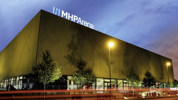 Die MHP Arena in Ludwigsburg bei Dämmerung, beleuchtet mit Straßenlaternen und umgeben von Bäumen. Der Himmel ist blau und rosa gefärbt., © MHPArena Ludwigsburg