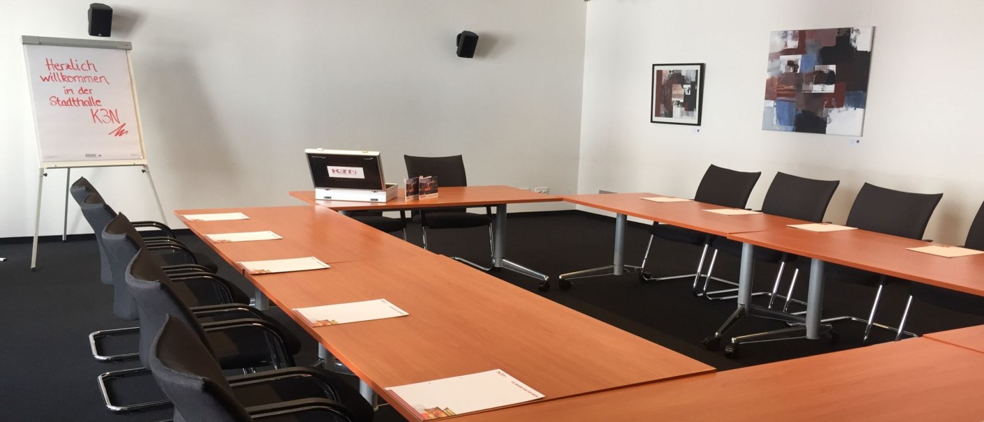 Konferenzraum mit U-förmigem Tisch, Stühlen und Flipchart. An der Wand hängen zwei abstrakte Bilder. Ein Projektor ist an der Decke montiert., © k3nmaisch_tf