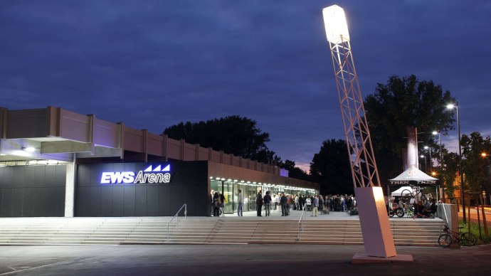 The EWS Arena at dusk, people standing in front of the entrance. An illuminated pole and a bicycle stand are visible., © Dietmar Strauss