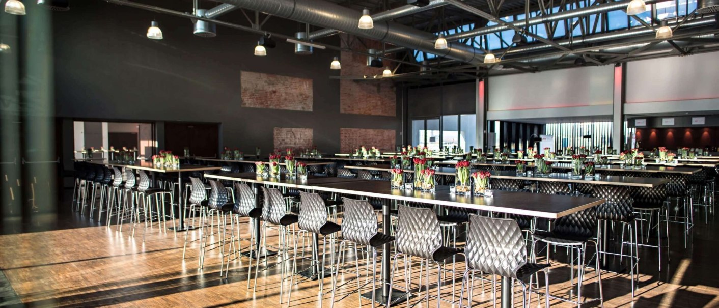 Modern hall with high tables and chairs, decorated with flowers. Industrial design with visible ventilation pipes and wooden floor., © rauschenbergercatering