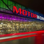 Motorworld Region Stuttgart at night, with illuminated lettering and a colorful glass front. Cars drive past and leave traces of light behind., © Alle Rechte bei Dieter Ege für EGEBILD. Vervielfältigungen, Veröffentlichungen, Drucke auch auszugsweise, sind nur gestattet mit ausdrücklicher schriftlicher Genehmigung von EGEBILD.