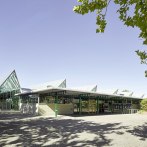 The Glaspalast Sindelfingen with its striking glass roof and green structures, surrounded by trees and a clear sky., © Glaspalast Sindelfingen