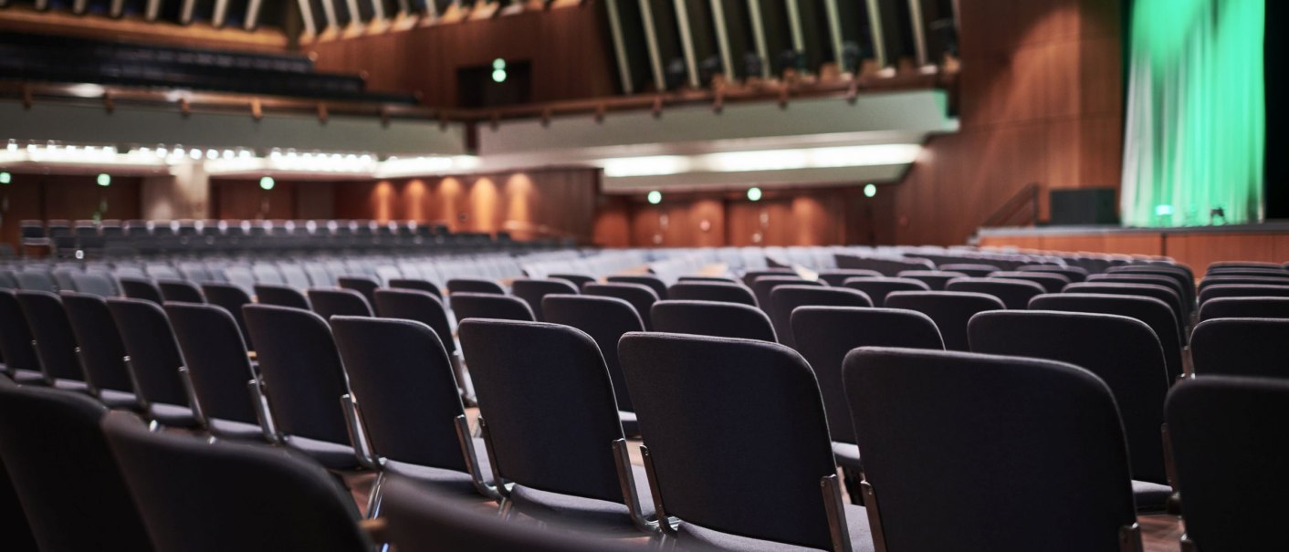 Empty hall with rows of chairs and stage in the Schwabenlandhalle Fellbach. Wooden walls and green lighting on the stage., © Peter Hartung Empty hall with rows of chairs and stage in the Schwabenlandhalle Fellbach. Wooden walls and green lighting on the stage., © Peter Hartung