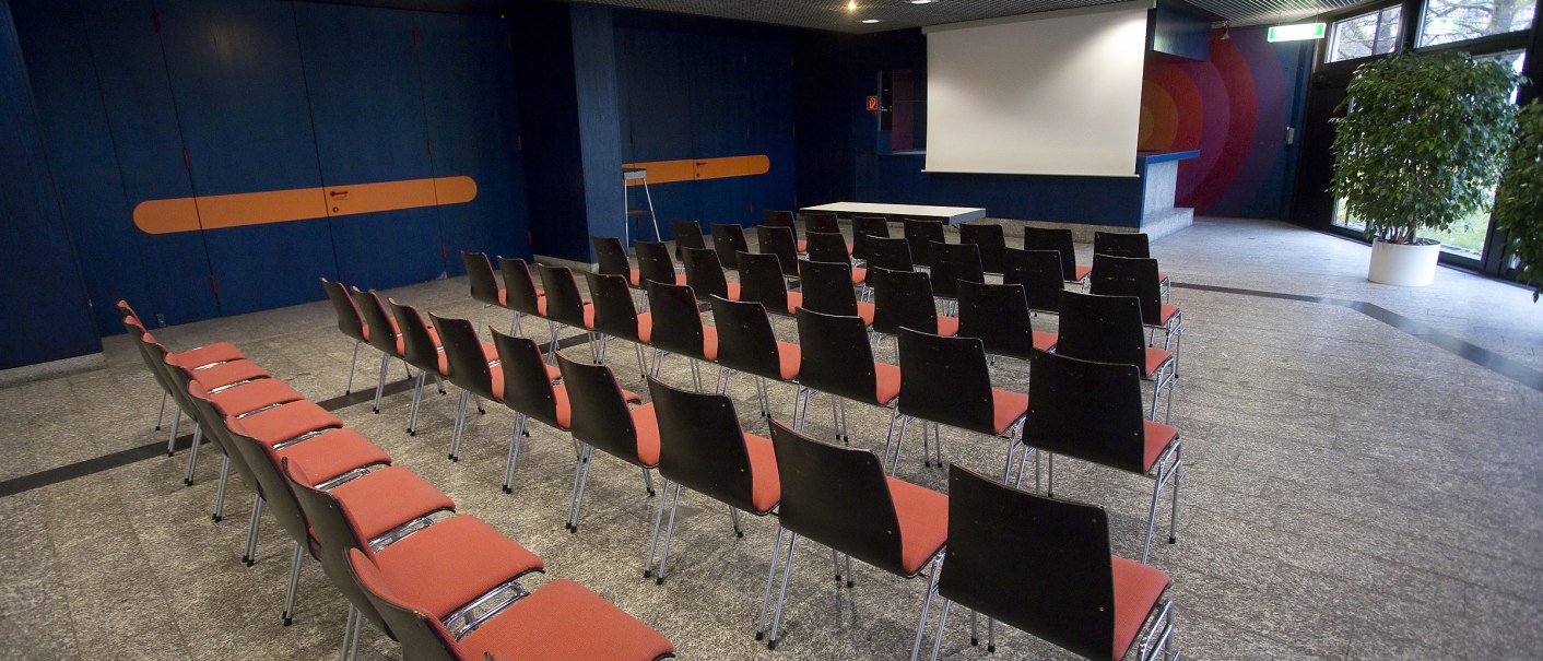 Ein leerer Konferenzraum mit roten Stühlen, einer Leinwand und blauen Wänden. Der Boden ist aus grauem Stein, und es gibt eine Pflanze in der Ecke., © eslive