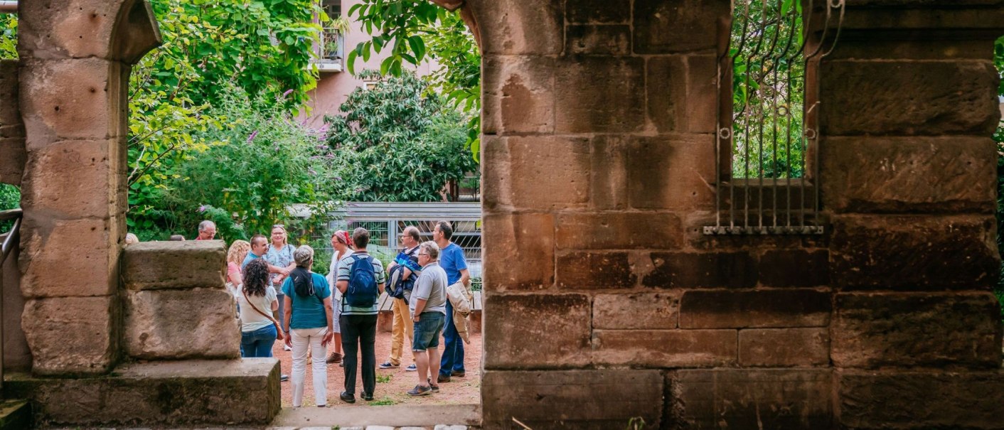 Eine Gruppe von Menschen steht in einem begrünten Innenhof, umgeben von alten Steinmauern. Im Hintergrund sind Pflanzen und ein Gebäude zu sehen., © Thomas Niedermüller
