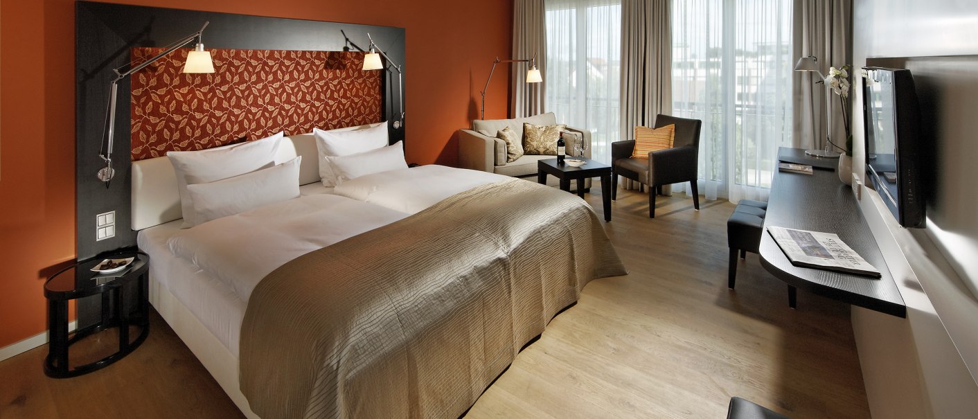 Ein modernes Hotelzimmer mit einem gro&szlig;en Doppelbett, einem Sofa, einem Schreibtisch und einem Flachbildfernseher. Die W&auml;nde sind orange gestrichen., &copy; Parkhotel Stuttgart Messe-Airport GmbH & Co KG