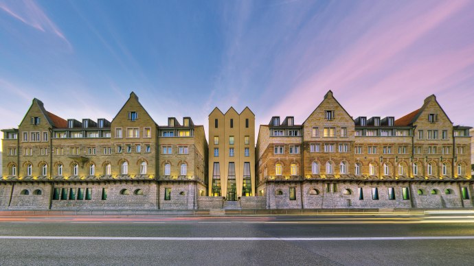 Das Arcotel Camino zeigt eine symmetrische Fassade aus Stein mit modernen Elementen, beleuchtet bei Sonnenuntergang. Eine Straße verläuft davor., © ARCOTEL Hotels