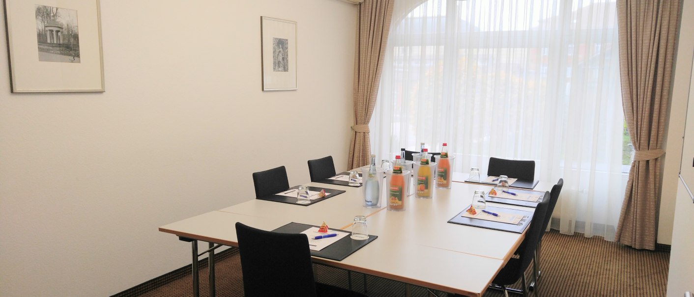 Heller Konferenzraum mit großem Tisch, sechs Stühlen, Getränken und Schreibmaterialien. Große Fenster mit Vorhängen, Bilder an der Wand., © nestor Hotel Ludwigsburg