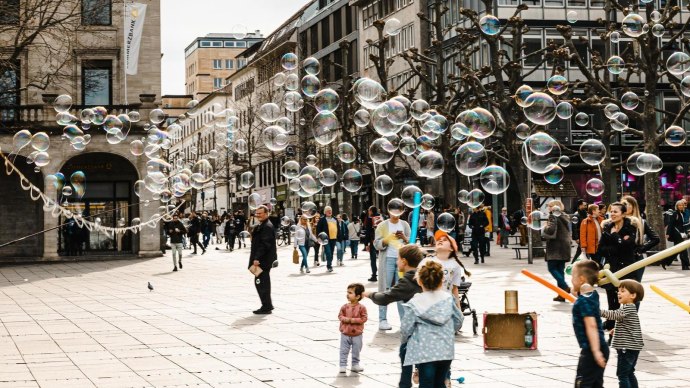 Ein Straßenkünstler erzeugt Seifenblasen auf einer belebten Straße. Kinder spielen begeistert mit den Blasen, während Passanten zuschauen., © Stuttgart-Marketing GmbH, Sarah Schmid Ein Straßenkünstler erzeugt Seifenblasen auf einer belebten Straße. Kinder spielen begeistert mit den Blasen, während Passanten zuschauen., © Stuttgart-Marketing GmbH, Sarah Schmid