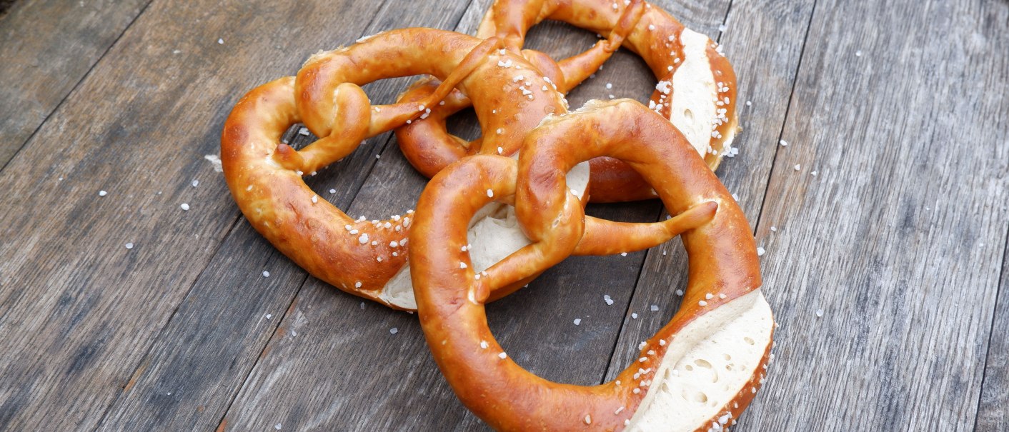 Drei frische Brezeln mit grobem Salz liegen auf einem rustikalen Holztisch., © Stuttgart Marketing GmbH