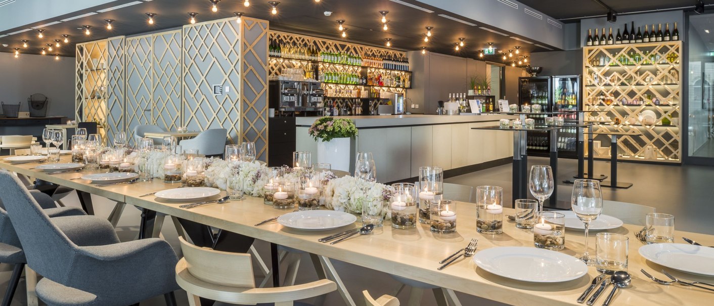 Elegant dining area with table setting, candles and flowers. In the background, a stylish bar with bottle racks and modern lighting., © Design Offices GmbH