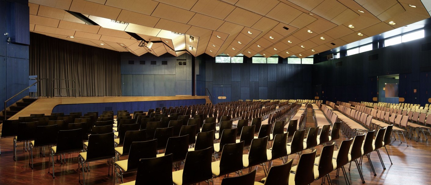 Großer Saal mit Holzfußboden, vielen Stuhlreihen und einer Bühne. Die Decke ist modern gestaltet, Fenster lassen Tageslicht herein., © eslive Großer Saal mit Holzfußboden, vielen Stuhlreihen und einer Bühne. Die Decke ist modern gestaltet, Fenster lassen Tageslicht herein., © eslive