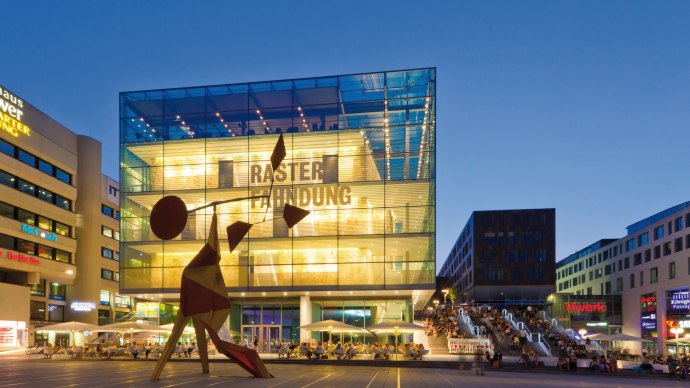 Das Kunstmuseum Stuttgart bei Abenddämmerung, beleuchtet mit moderner Skulptur im Vordergrund und Menschen auf der Treppe daneben., © SMG, Werner Dieterich