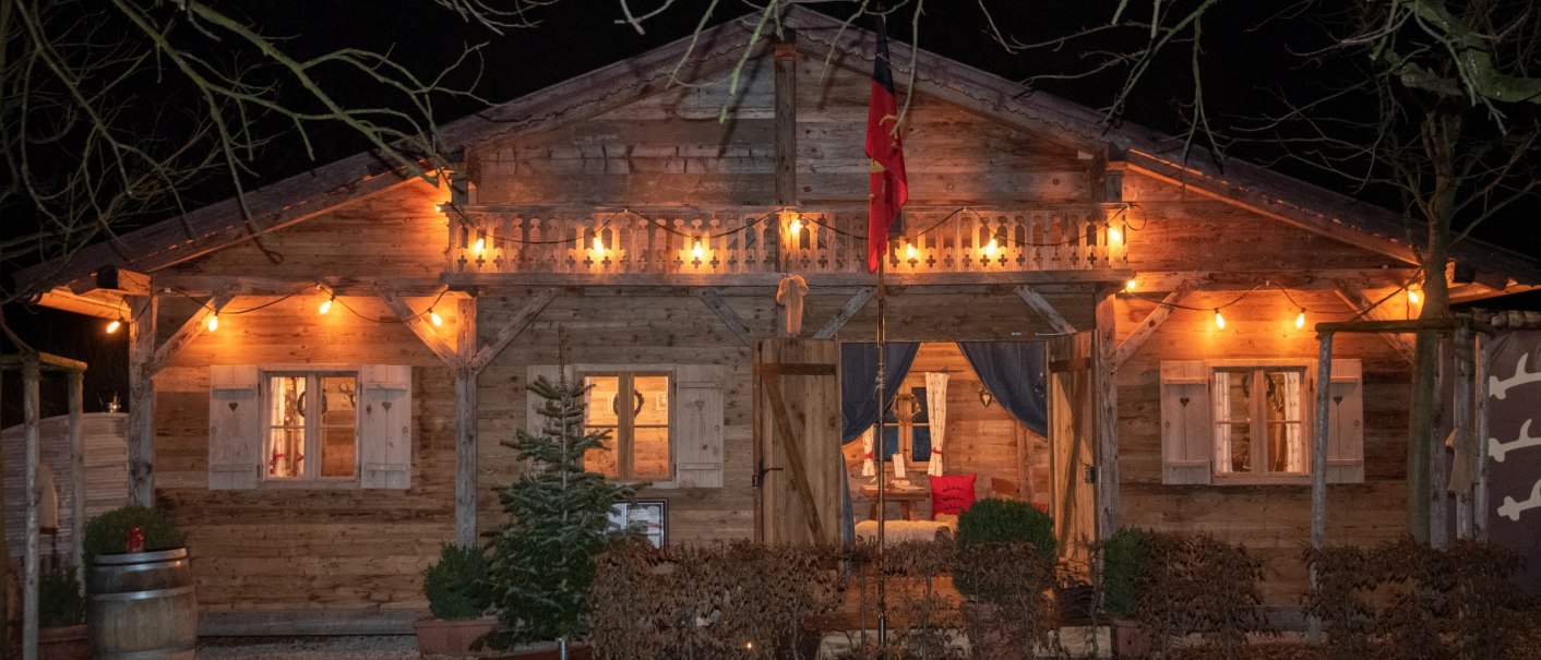 A cozy, illuminated wooden hut at night, surrounded by trees and plants. There is a Christmas tree and a wine barrel in front of the hut., © atelier-fuessinger A cozy, illuminated wooden hut at night, surrounded by trees and plants. There is a Christmas tree and a wine barrel in front of the hut., © atelier-fuessinger