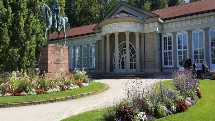 Der Kursaal Bad Cannstatt mit klassischer Fassade, Reiterstatue und bunten Blumenbeeten im Vordergrund an einem sonnigen Tag., © smg