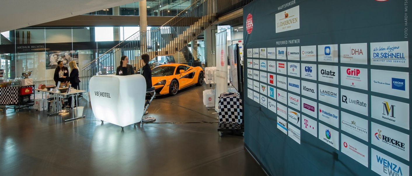Foyer of the V8 Hotel with reception desk, people in conversation, a sports car and a sponsor board., &copy; Hardy Mutschler