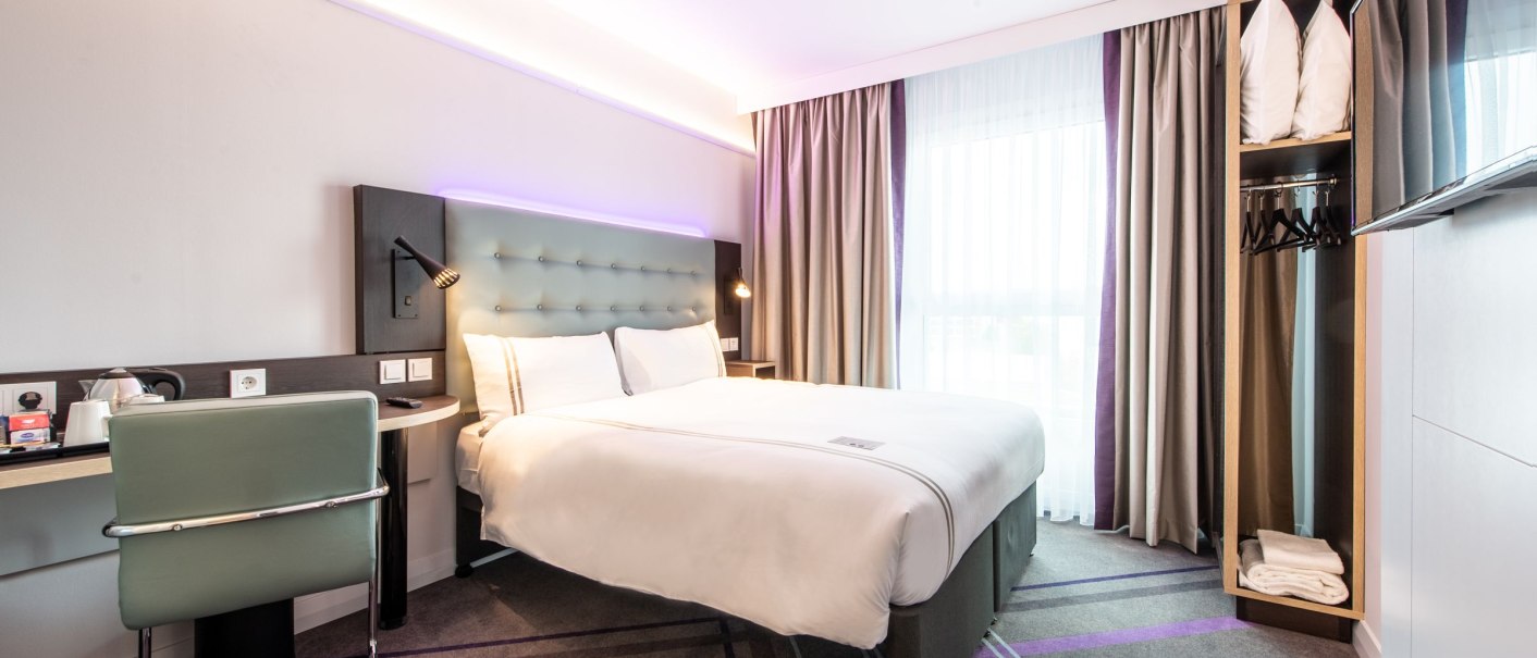 Modernes Hotelzimmer mit Doppelbett, Schreibtisch, Stuhl, Kleiderschrank und großem Fenster mit Vorhängen. Helle und stilvolle Einrichtung., © Guenter Lenz