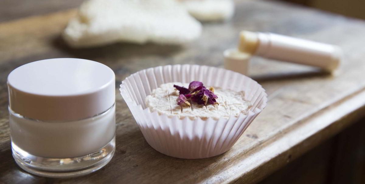 Ein Glas mit Creme, ein festes Kosmetikprodukt in einem Papierförmchen und ein Lippenpflegestift auf einem Holztisch., © hoponopo GmbH