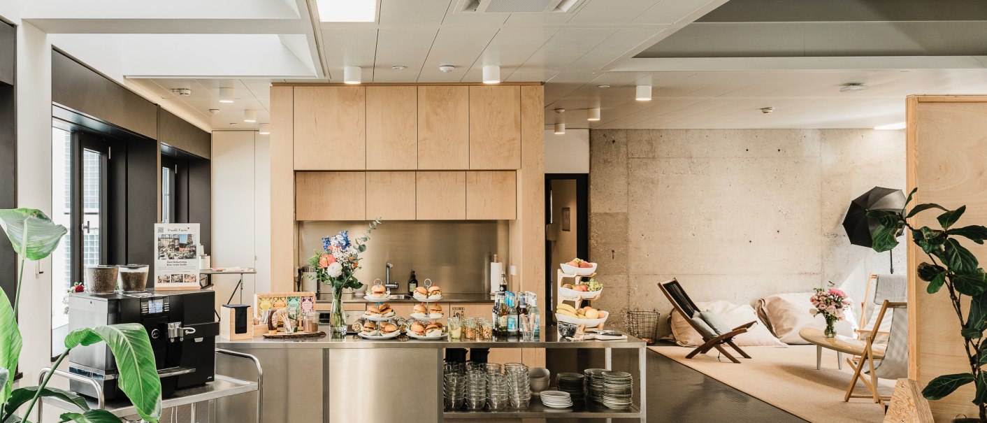 Modern kitchen with wooden cupboards, stainless steel counter and coffee machine. A cozy seating area with plants and decorations in the background., © OutOfOffice GmbH