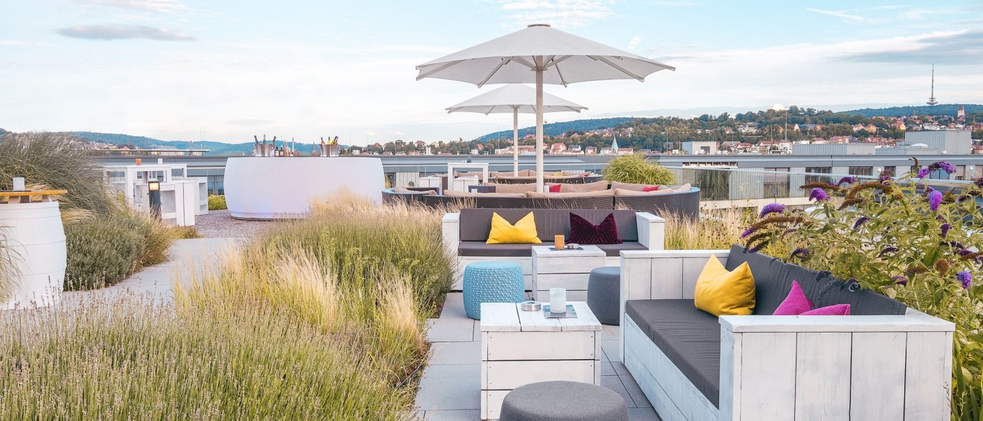 Modern roof terrace with white seating furniture, colorful cushions and parasols. Plants and city views in the background under a blue sky., &copy; JazHotels