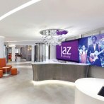 Moderne Hotelrezeption mit geschwungenem Tresen, großem Bildschirm mit 'Jaz in the City' Logo und stilvoller Beleuchtung., © Jaz Hotel Stuttgart
