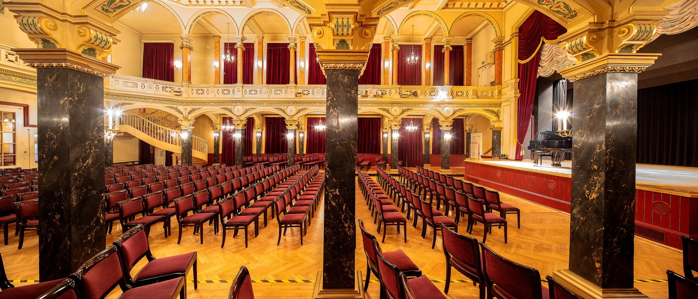 Magnificent concert hall with red chairs, golden decorations and a grand piano on the stage. Elegant architecture with columns and chandeliers., © Stauch