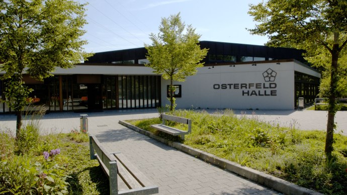 Die Osterfeldhalle von außen, umgeben von Bäumen und Pflanzen. Ein gepflasterter Weg führt zu Bänken vor dem Gebäude., © eslive