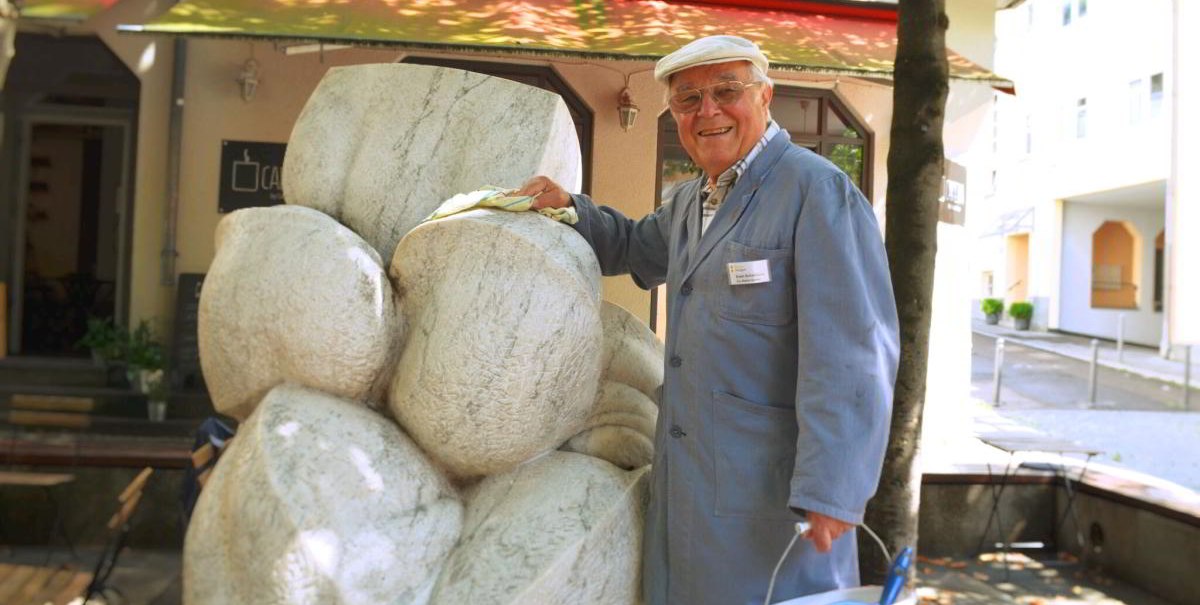 Ein älterer Mann in blauer Arbeitskleidung reinigt eine Steinskulptur. Er trägt eine Mütze und hält einen Eimer in der Hand. Im Hintergrund ist ein Gebäude., © Stuttgart-Marketing GmbH