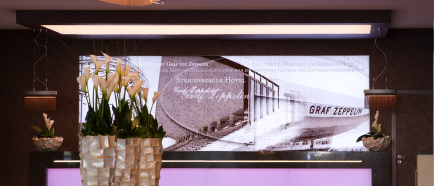 Elegant lobby of the Steigenberger Graf Zeppelin Hotel with stylish lighting, flower arrangements and a historical picture in the background., © Steigenberger Hotels AG