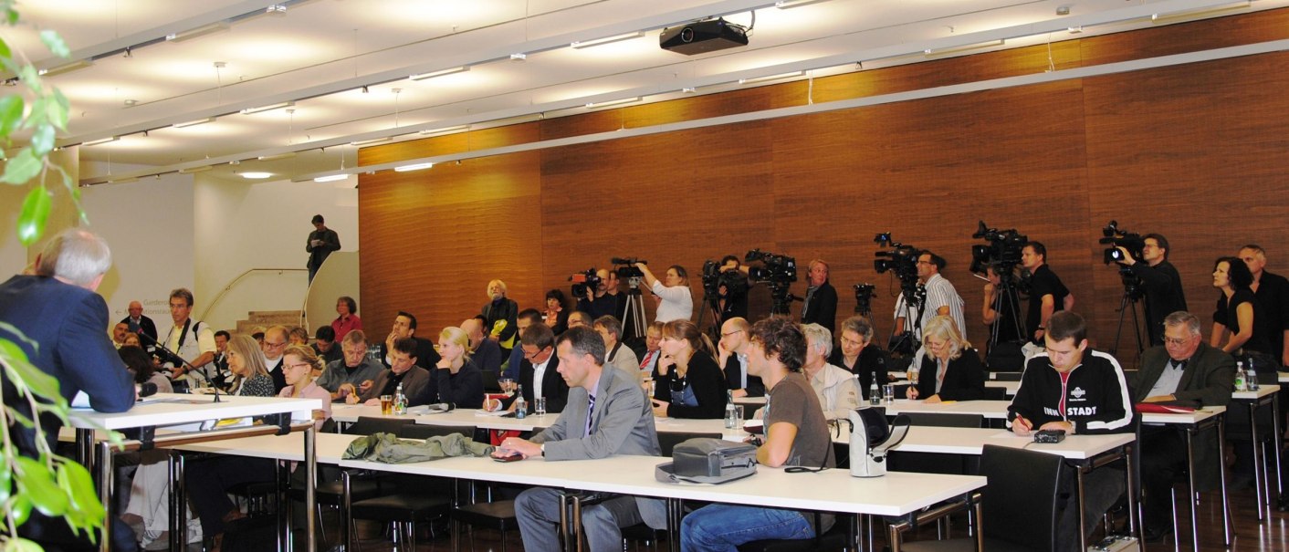 Ein Konferenzraum mit vielen Menschen, die an Tischen sitzen. Einige machen Notizen, während Kamerateams im Hintergrund filmen., © hdkk_gf