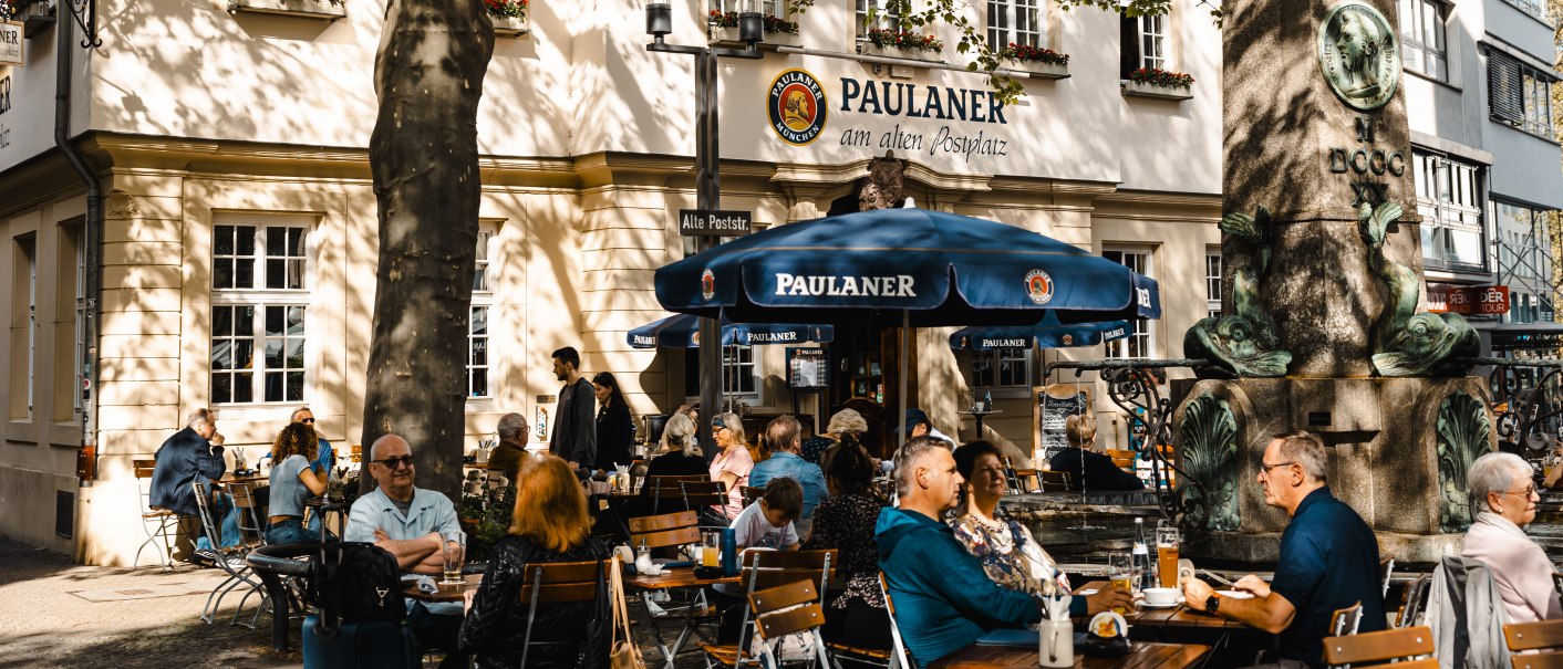Menschen sitzen im Außenbereich eines Paulaner-Restaurants. Ein großer Baum spendet Schatten, und ein Brunnen ist im Vordergrund zu sehen., © SMG Stuttgart Marketing GmbH - Sarah Schmid