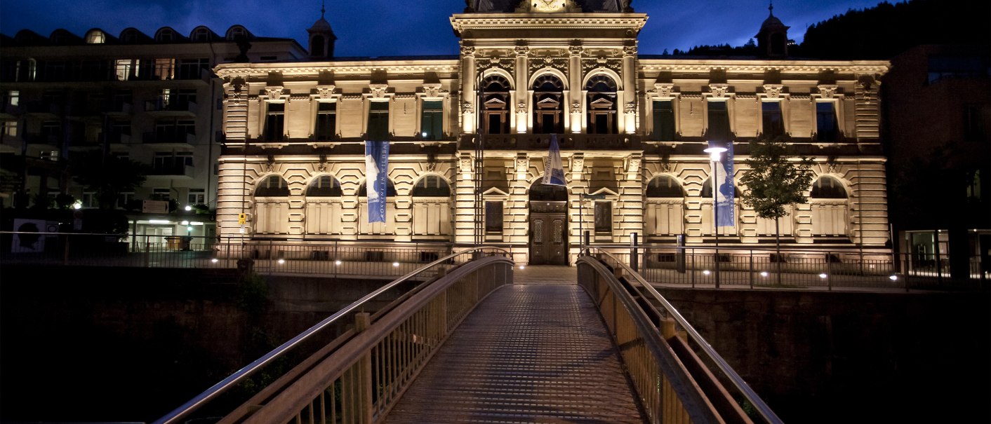 An illuminated building at night, the Forum König-Karls-Bad, with a bridge in the foreground. The sky is dark blue., © Forum Koenig-Karls-Bad An illuminated building at night, the Forum König-Karls-Bad, with a bridge in the foreground. The sky is dark blue., © Forum Koenig-Karls-Bad